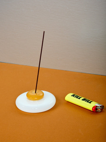 An egg shaped incense holder with incense next to a yellow lighter.