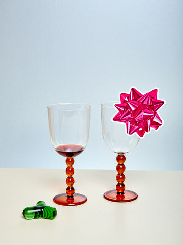 Two wine glasses with colorful stems, one filled with red wine, on a light surface with a light blue background.