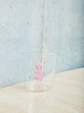 A small cup with a pink rabbit made of glass inside sits on a cream table. 