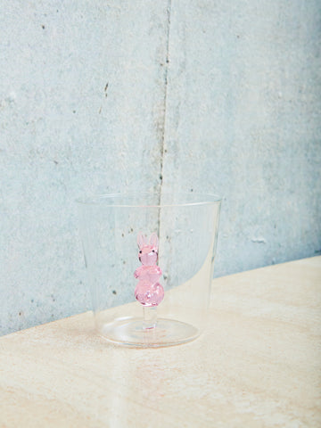 A small cup with a pink rabbit made of glass inside sits on a cream table. 