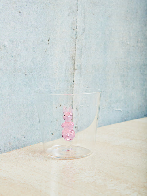 A small cup with a pink rabbit made of glass inside sits on a cream table. 