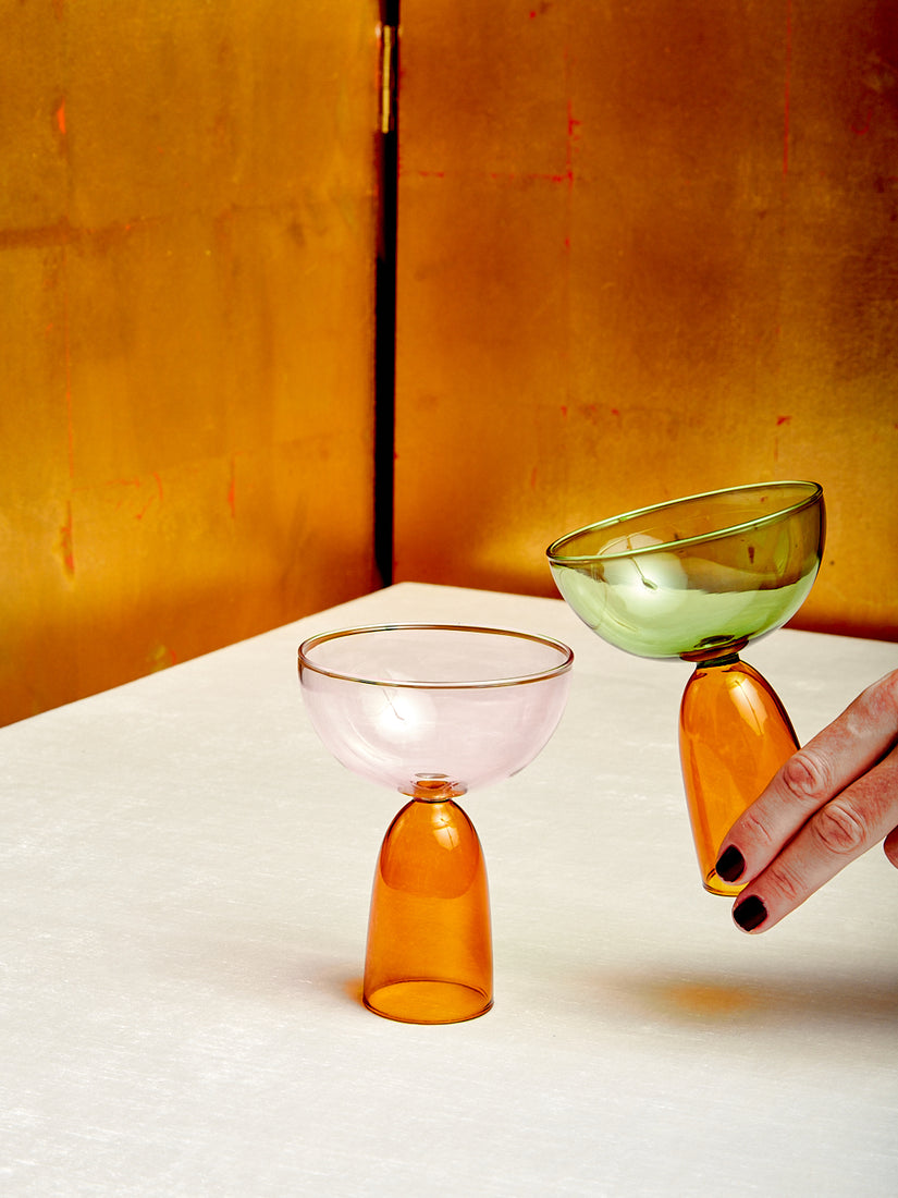 A pink/amber and green/amber coco coupe. A hand holds the green glass up at a slight angle towards the pink glass.