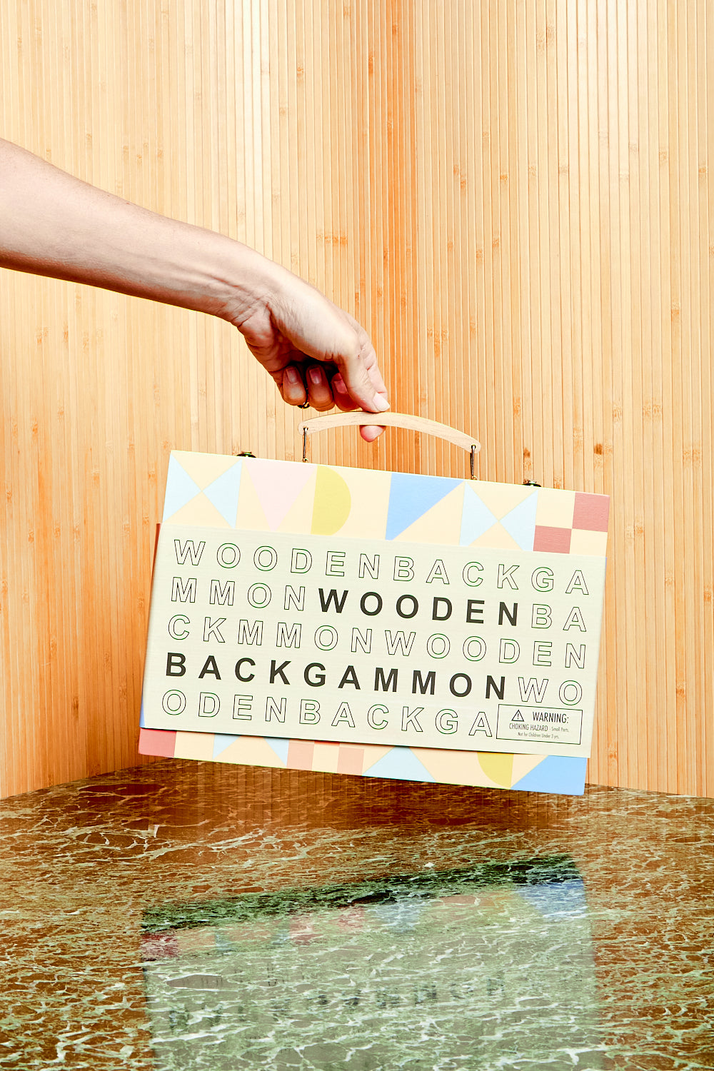 A hand holds Sunnylife's Wooden Backgammon game up by its handle. The game is in its branded graphic packaging sleeve.