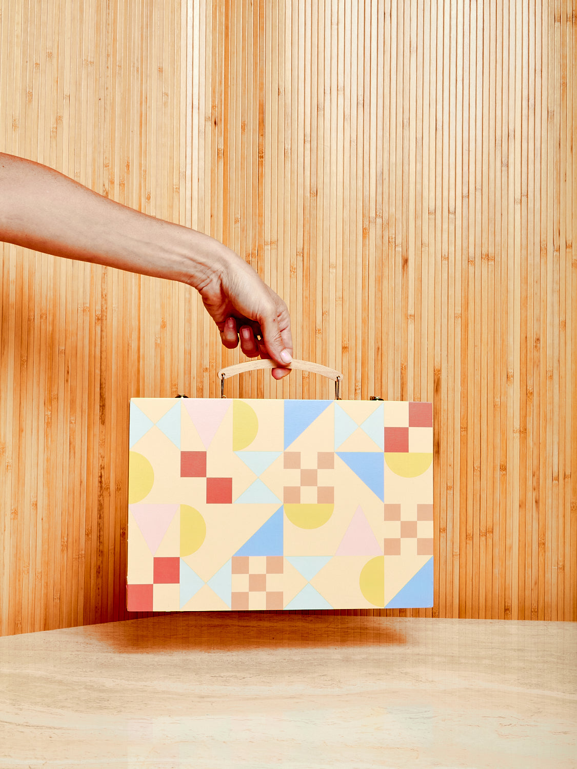 A hand holds up the Sunnylife Travel Backgammon by its handle displaying its colorful  and geometric exterior.