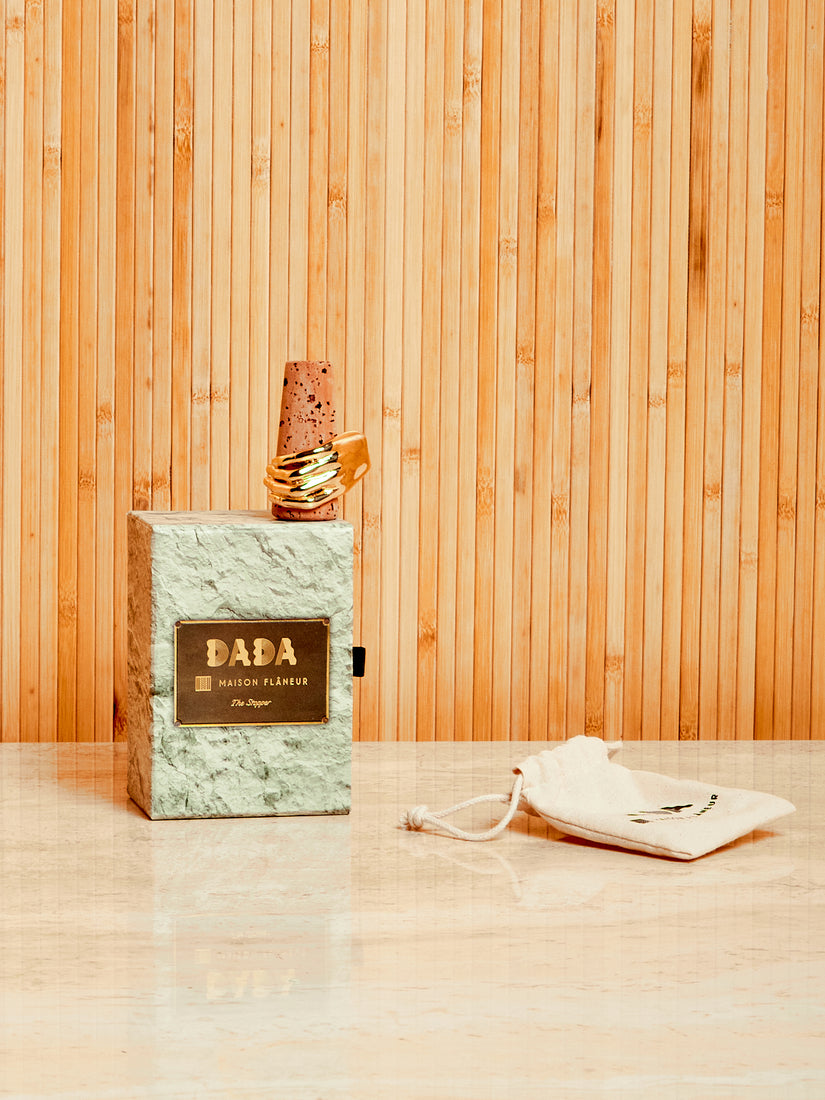 The Dada cork bottle stopper with brass hand sits atop its box next to the canvas pouch it comes in.