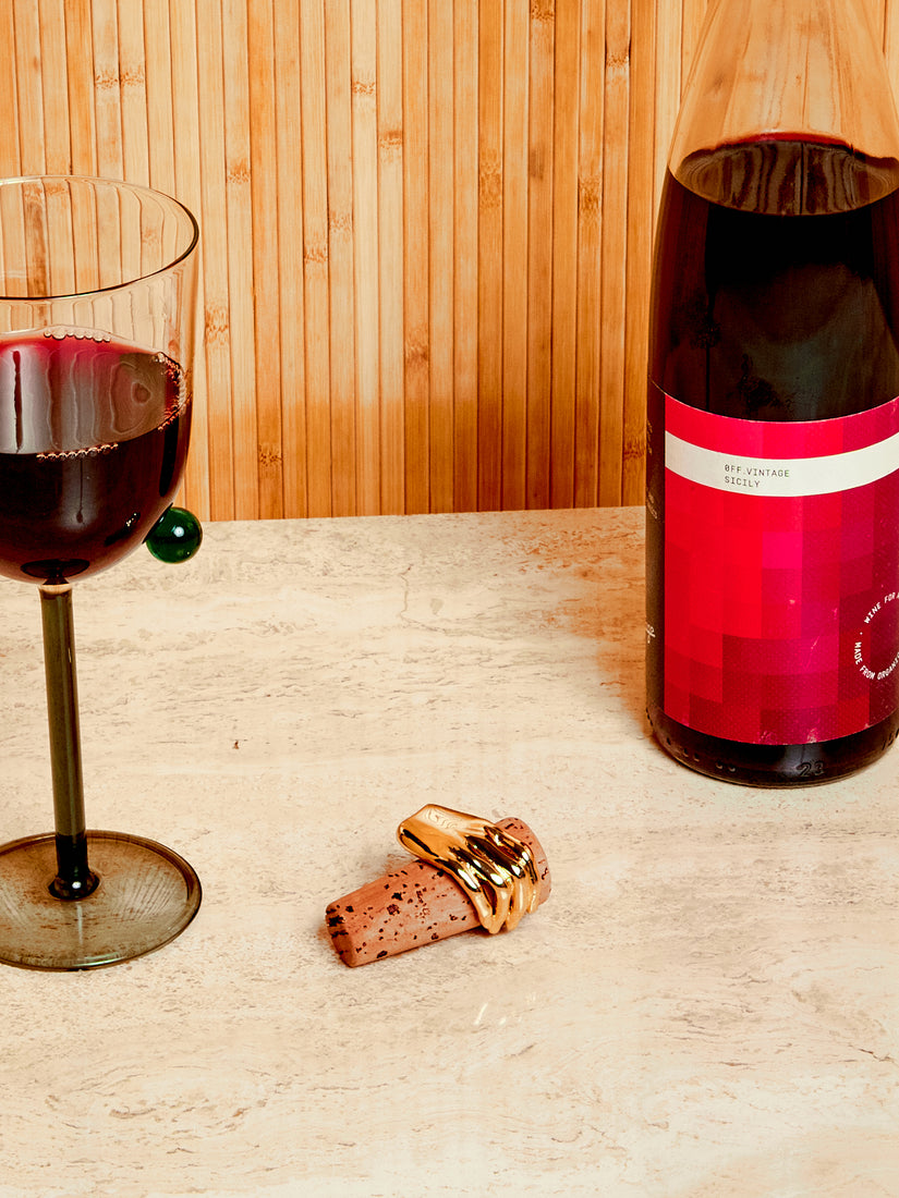 A Pompom wine glass, bottle of wine, and wine bottle stopper.