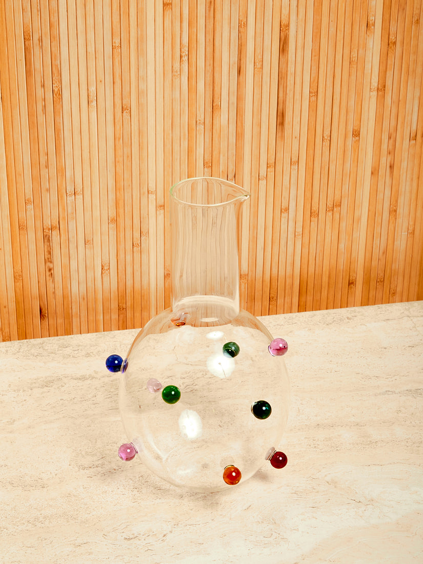 A clear carafe with a straight neck, spouted lip, and rounded body featuring multicolored glass dots.