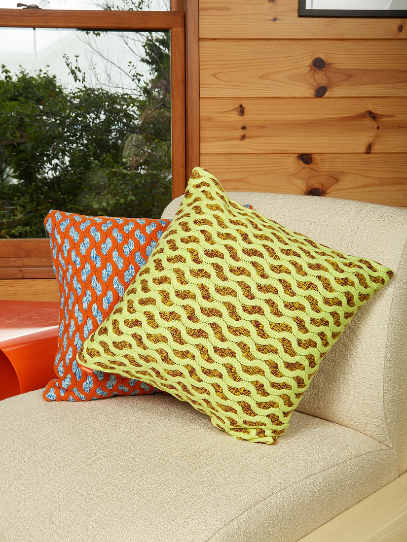 Two Squiggle Cable Pillows by Verloop in orange and lime sitting in a wooden room on a cream Uma T4 chair.