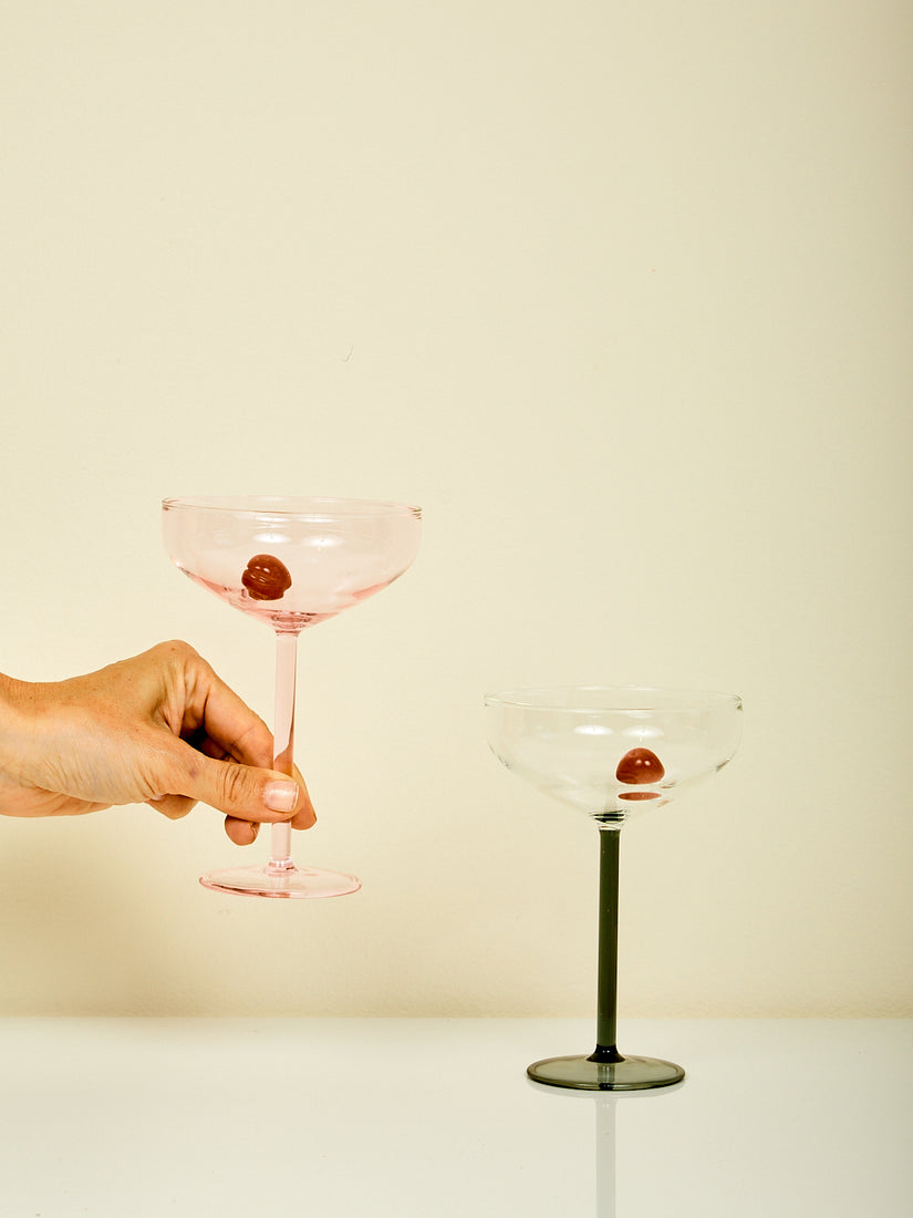 Manhattan Coupe Glasses by Maison Balzac. A hand hold the pink colorway while the smoke colorway rests on a surface.