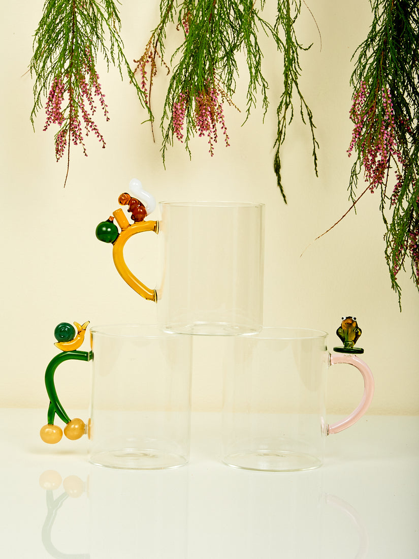 Three glass animal farm mugs by Ichendorf Milano. Each mug features a unique colored handle and a squirrel, snail, or frog adornment. The squirrel mug rests atop the lips of the snail and frog mugs.