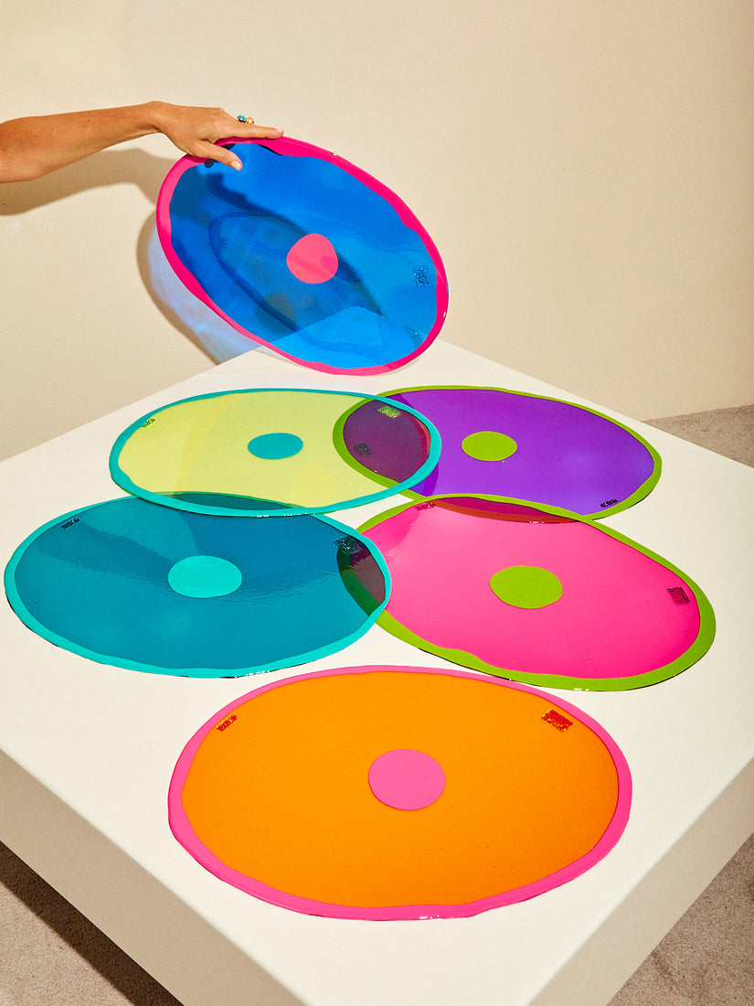 Six different colored Dot Table-Mates Placemats by Gaetano Pesce for Fish Design on a square white tabletop. A hand lift ups the blue/fuschia placemat.