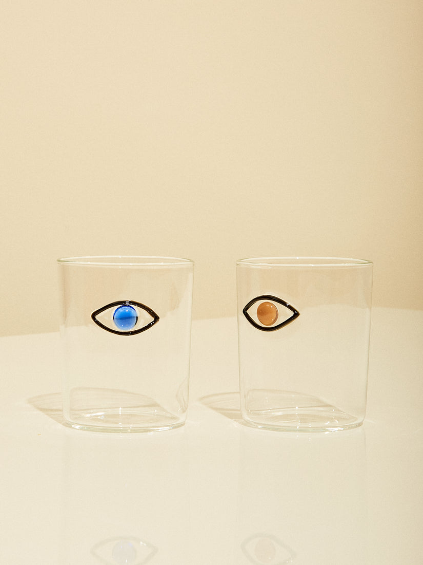 A pair of glass cups with flame-worked eye design. The left glass features a blue eye and the right glass has a light brown eye.