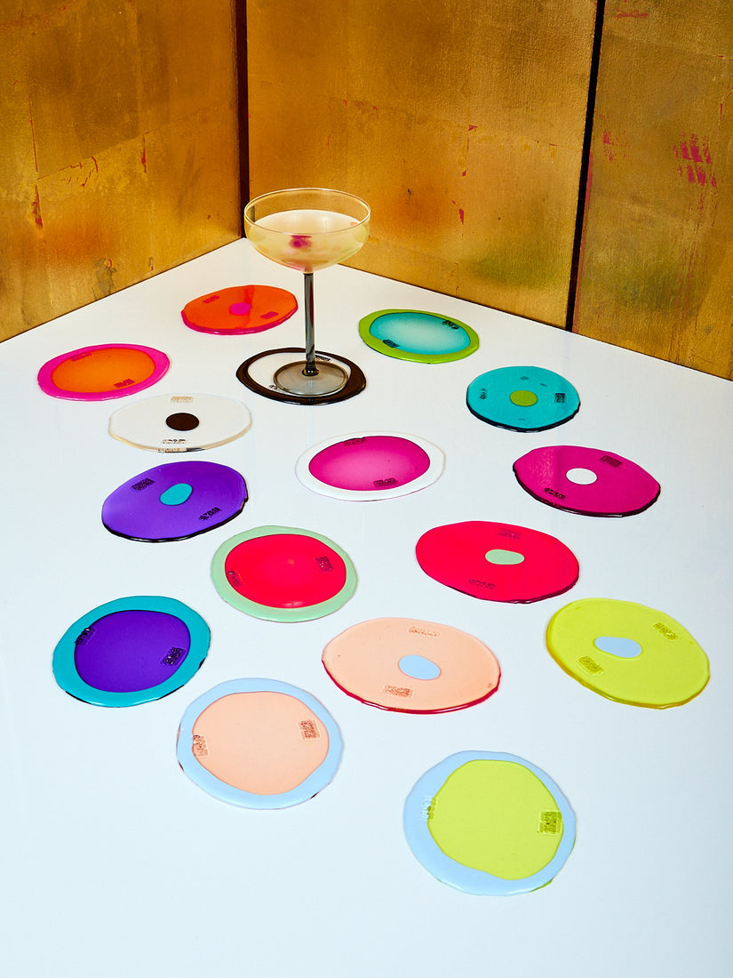 Eight different colorways of the Set of 2 Table-Mates resin coasters by Gaetano Pesce for Fish Design scattered across a white table top. One coaster has a Manhattan Glass by Maison Balzac.