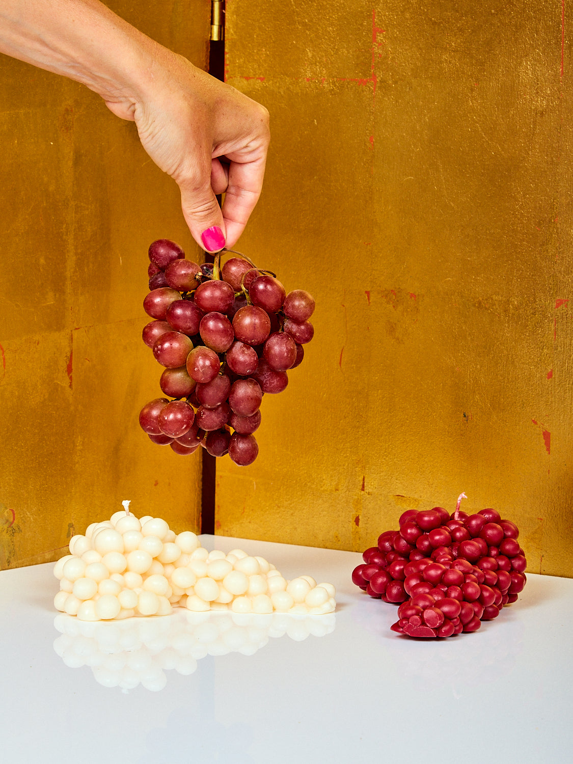 A hand holds a bunch of grapes over a white and wine Grape Candle by Maison Balzac.