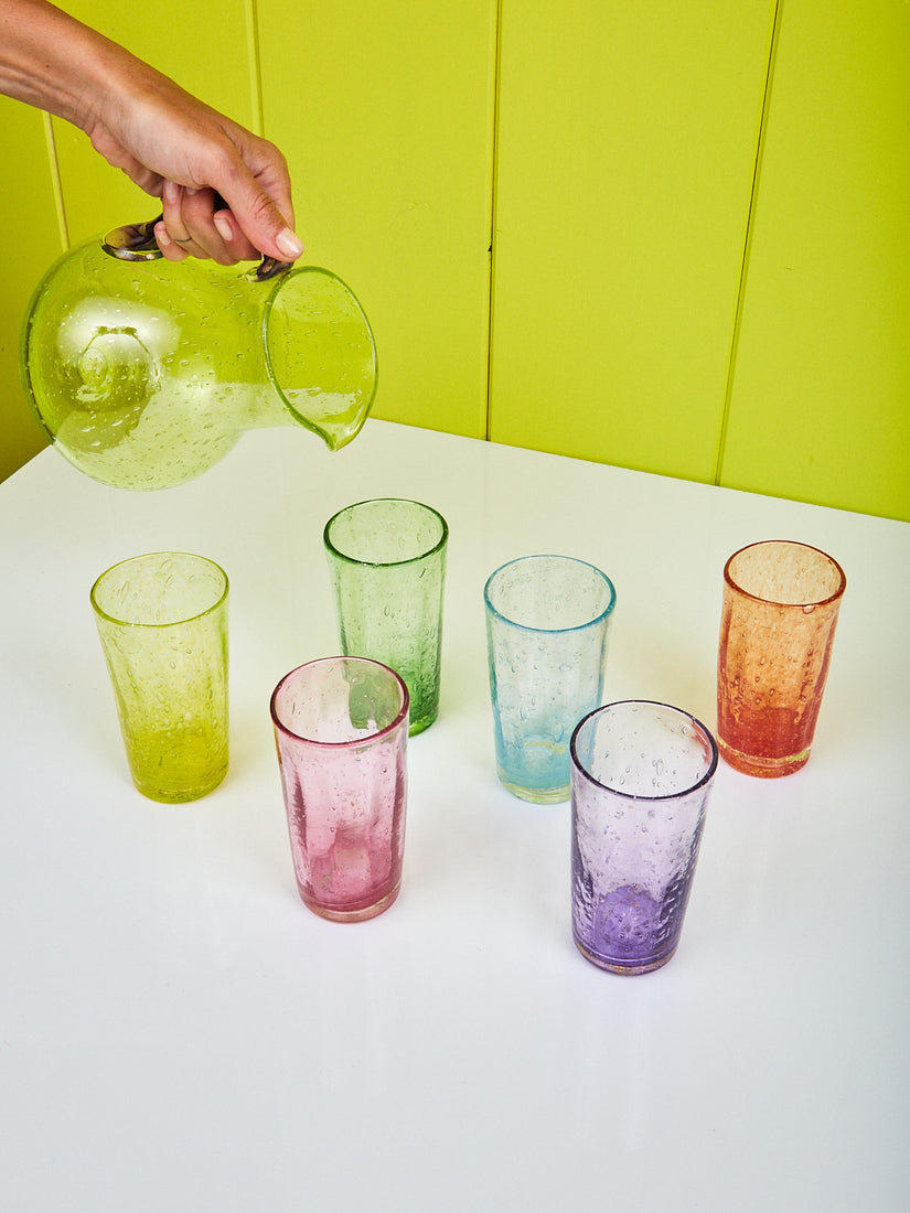A hand holds up the Bubble Glass Jug over six Orangeade Glasses in different colors.