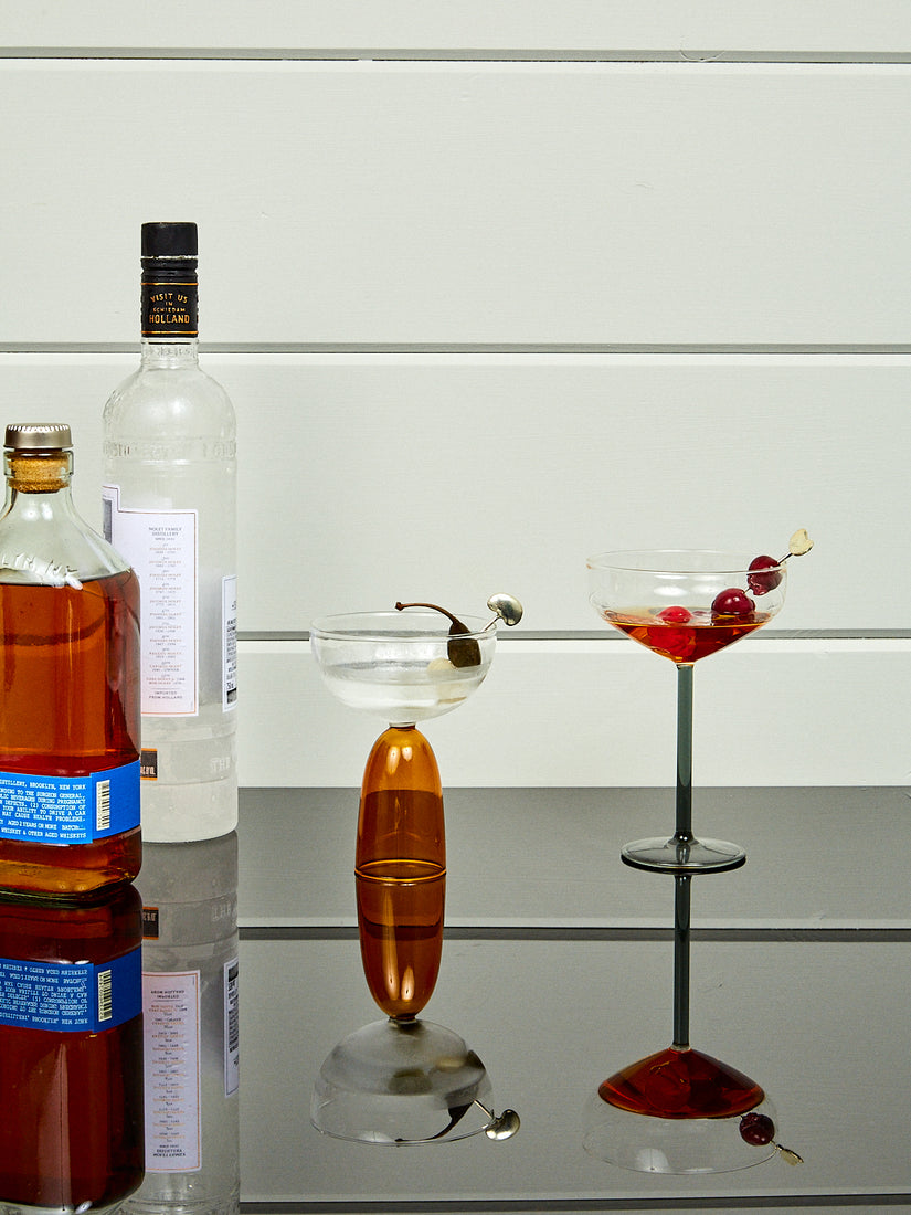 Two cocktails utilizing Bean Toothpicks next to bottles of spirits.