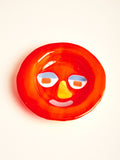 A bright red bowl with abstract face featuring a yellow nose, and pink mouth.