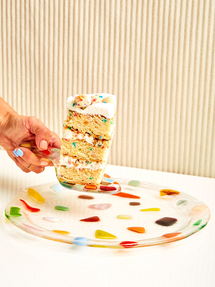 A hand holds up a glass confetti cake server with a rainbow slice of cake on it, hovering above a confetti plate.