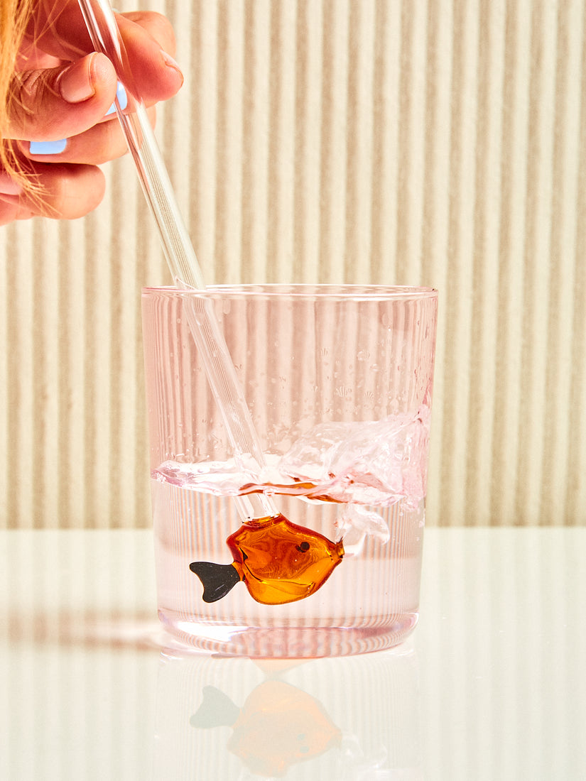 Some blows bubbles into a pink glass of water through an amber fish straw by Ichendorf Milano.