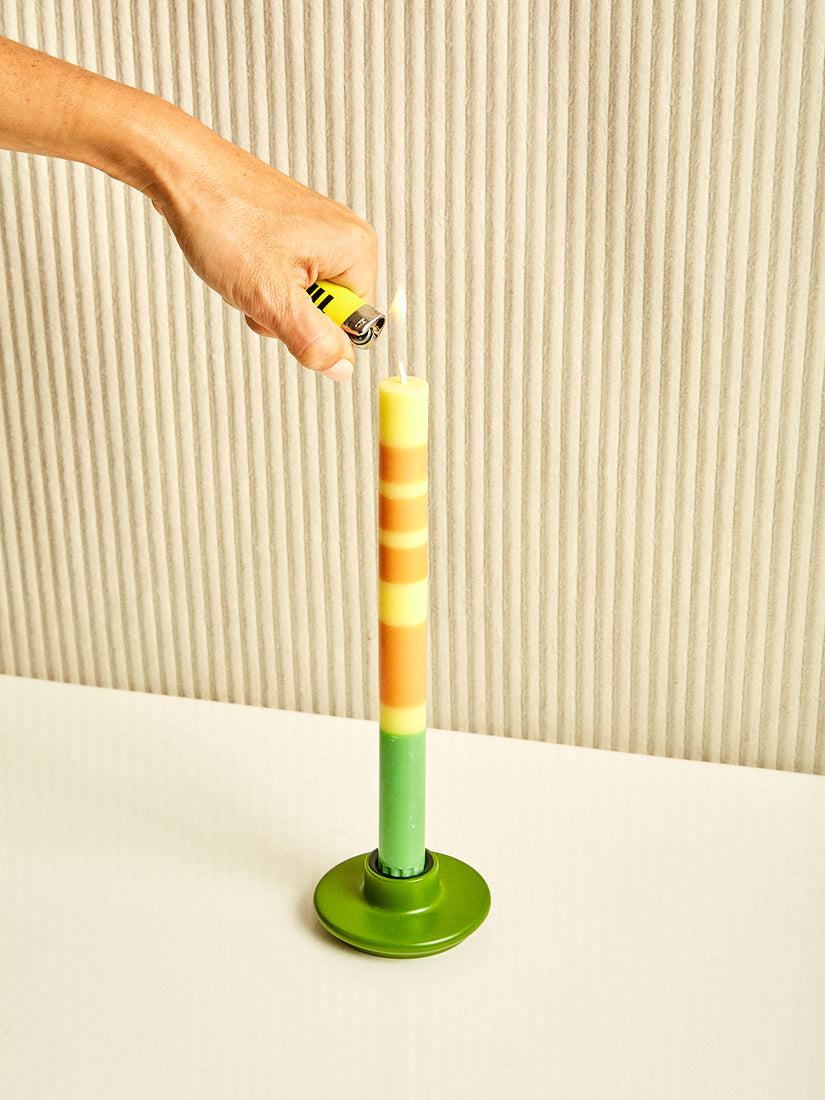 A hand lights a yellow, orange, and green taper candle sitting in an olive green candle holder by British Colour Standard.