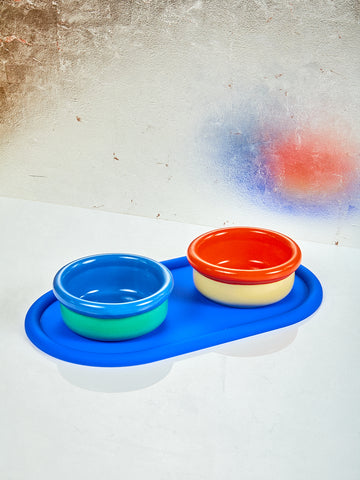 A blue and green bowl and a red and yellow bowl sit on a blue silicone mat. 