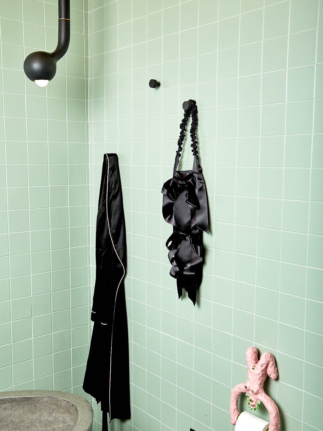 A black baguette bag hangs in a sage green tiled bathroom.