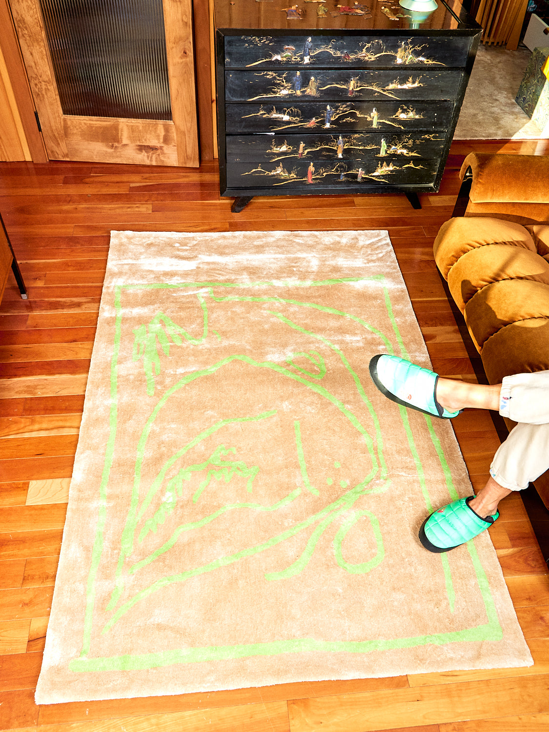 Freya, in Gold rug by Cold Picnic lays on wooden floor. A persons feet show with green slippers on.  