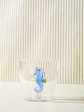 A small cup with a blue seahorse made of glass inside it sits on a cream table.