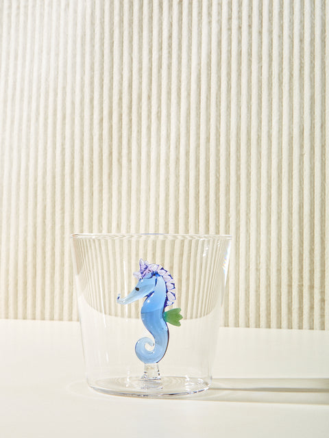 A small cup with a blue seahorse made of glass inside it sits on a cream table.