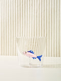 A small cup with a sardine figure made of glass inside sits on a cream table. The sardine is light pink and blue.