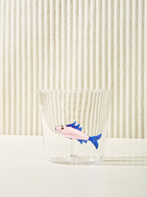 A small cup with a sardine figure made of glass inside sits on a cream table. The sardine is light pink and blue.