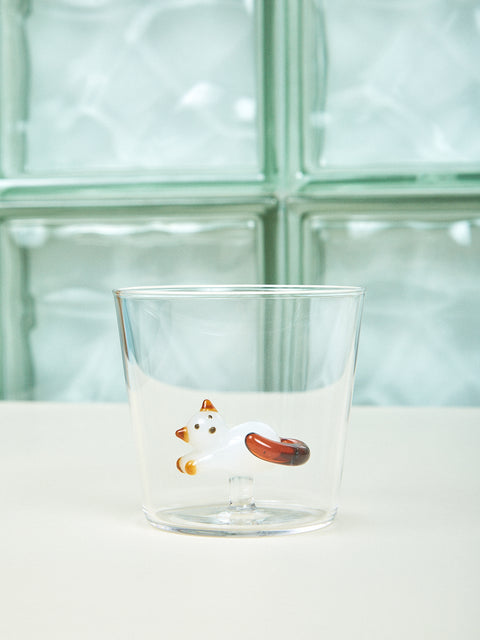 A small cup with a white and brown cat made of glass inside it sits on a cream table. 