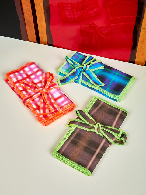 Three different set of napkins lay on top of a white table. One set of napkins is pink and orange, the second set is brown and lime green, and the last set is dark blue and purple. 