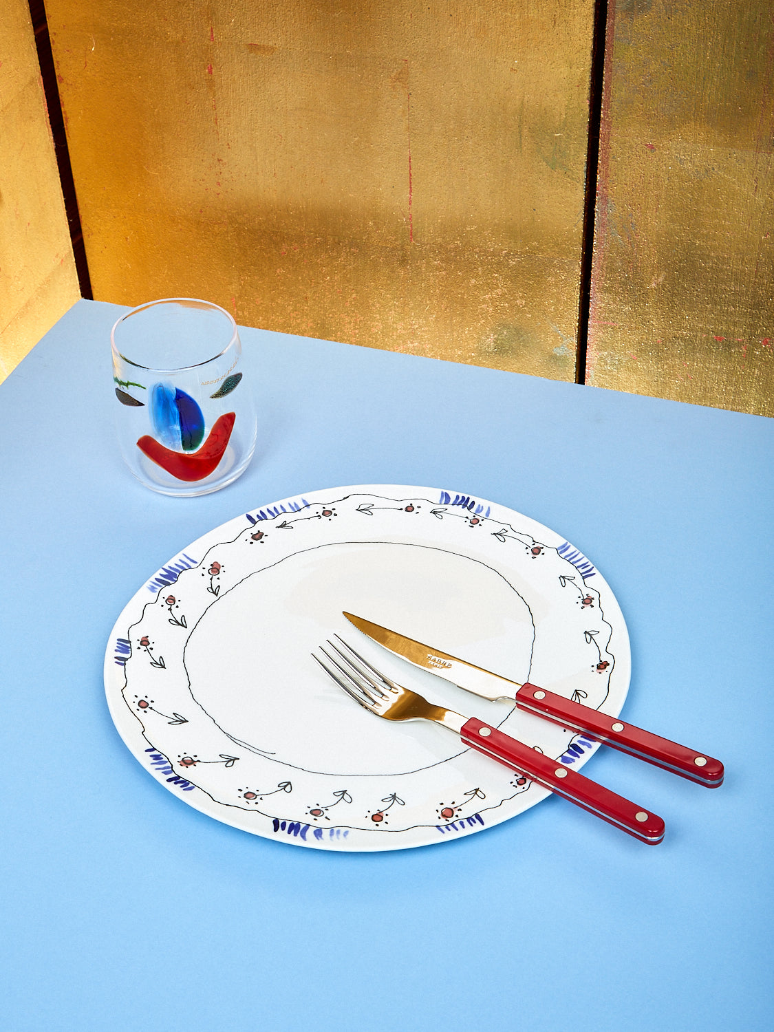 A plate by Marni lay on top of a blue table. On the plate there is a red fork and knife. On the top left there is a cup with a face shape on it.