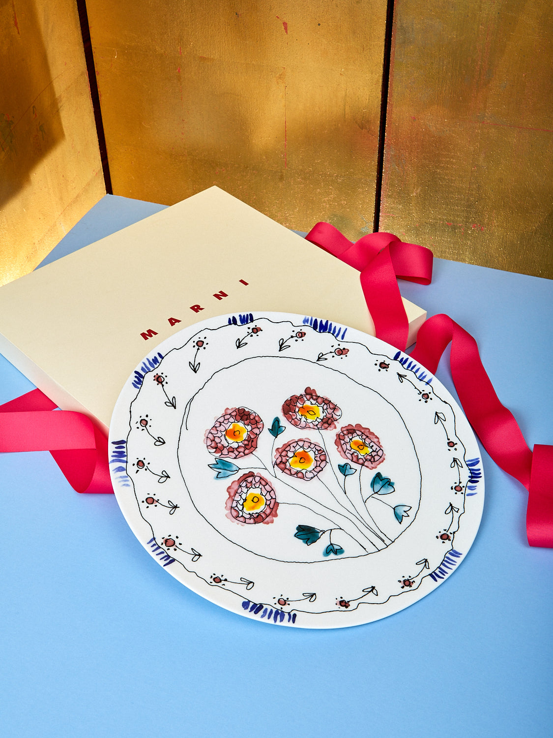 Marni serving plate sits on top of light blue table giving a good view of the stenciled flowers on the plate. An empty Marni box shows behind the plate with pink ribbon laid around it.