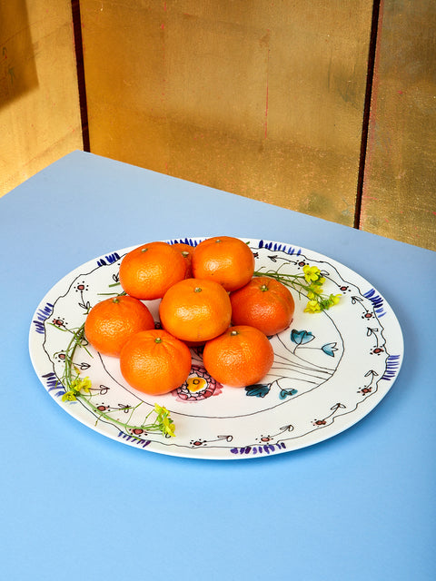 Marni serving plate sits on top of light blue table. On the Marni plate, a bundle of mandarins sit with tiny green and yellow flowers beside them. 