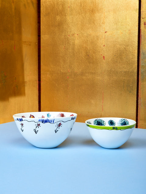 Two bowls by Marni lay on top of a blue table with a gold background. The bowl on the left side is medium sized and has white and magenta coloring. The bow on the right is small sized and has white and blue coloring.