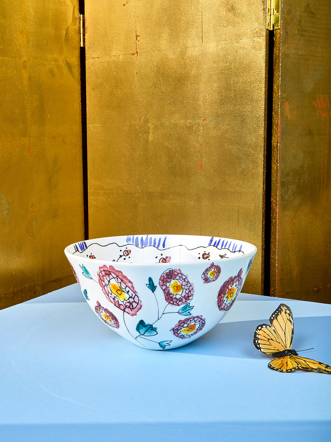 Large serving bowl by Marni sits on a light blue table. Side view showing the stencils of flowers. A fake butterfly sits on the left side of the table.