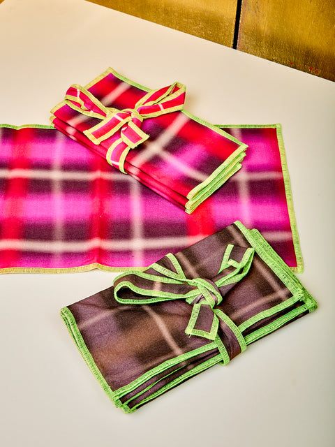 Napkins lay on top of a white table. One set of napkins has pink, brown and lime colors in a grid like shape. The other Napkin set has brown and lime green colors. Both are folded up with a bow wrapping them together.