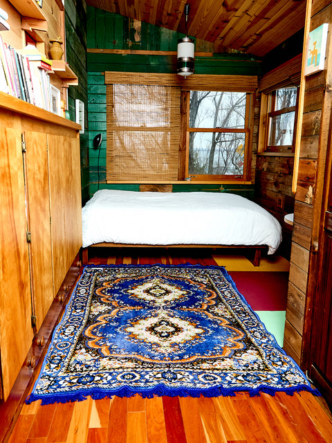 The Festival Rug in blue sits on a hardwood floor in a bedroom. The bed behind the rug has the Reversible blanket/rug displayed on top of it. The Blue Festival Rug has an intricate design.