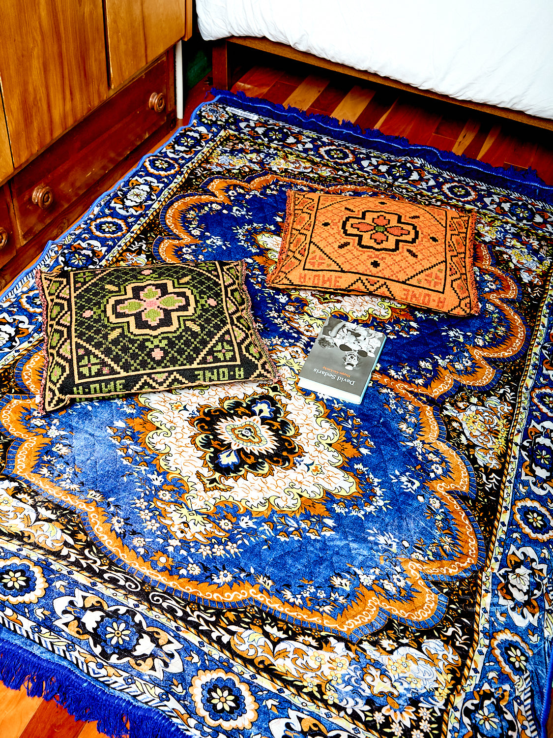 The Festival Rug in blue sits on a hardwood floor. The photo shows the rug zoomed in a bit more. The Woven Throw pillows by Peubco also sit on top of the rug. 