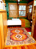 The Festival Rug in Gold/Orange sits on a hardwood floor in a bedroom. The bed behind the rug has the reversible blanket/rug displayed on top of it. The Gold Festival Rug has an intricate design. 