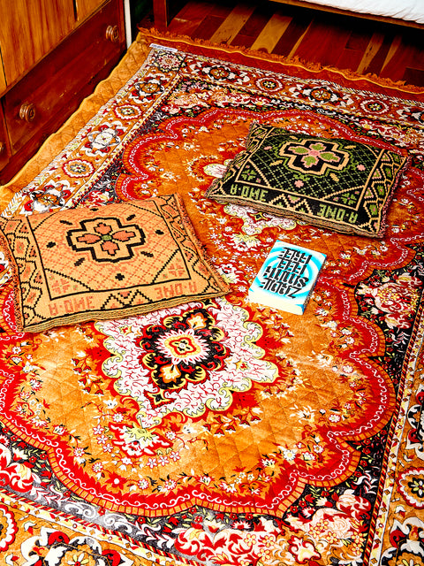 The Hybrid woven pillows in black/orange and black/green sit on top of a Gold Festive Rug by Puebco. There is a blue book sitting between the pillows.