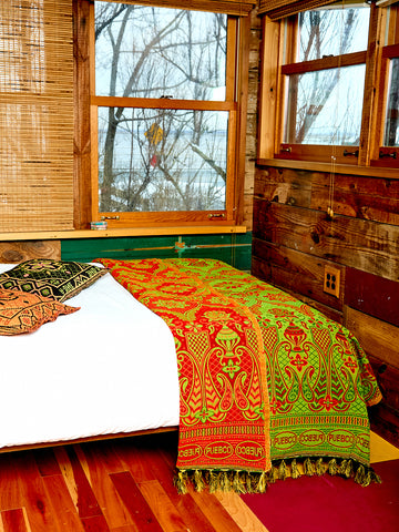 The Reversible blanket or rug sits on the egde of a bed in a room with hardwood floors and two windows. The blanket or rug is lime green and red. 