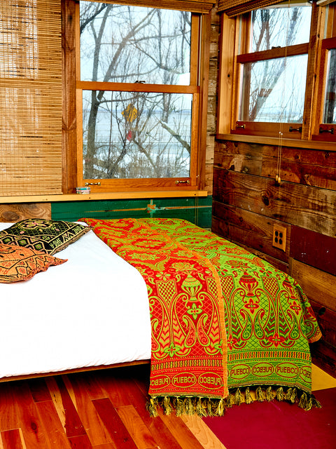 The Reversible blanket or rug sits on the egde of a bed in a room with hardwood floors and two windows. The blanket or rug is lime green and red. 