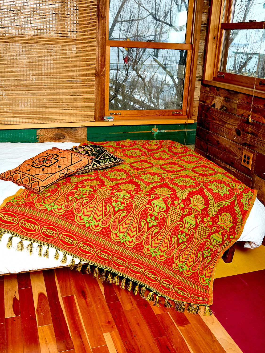 The Heavy Reversible Blanket/Rug lays on top of a bed. The Woven throw pillows are displayed with it. The blanket/rug is showing the red side.