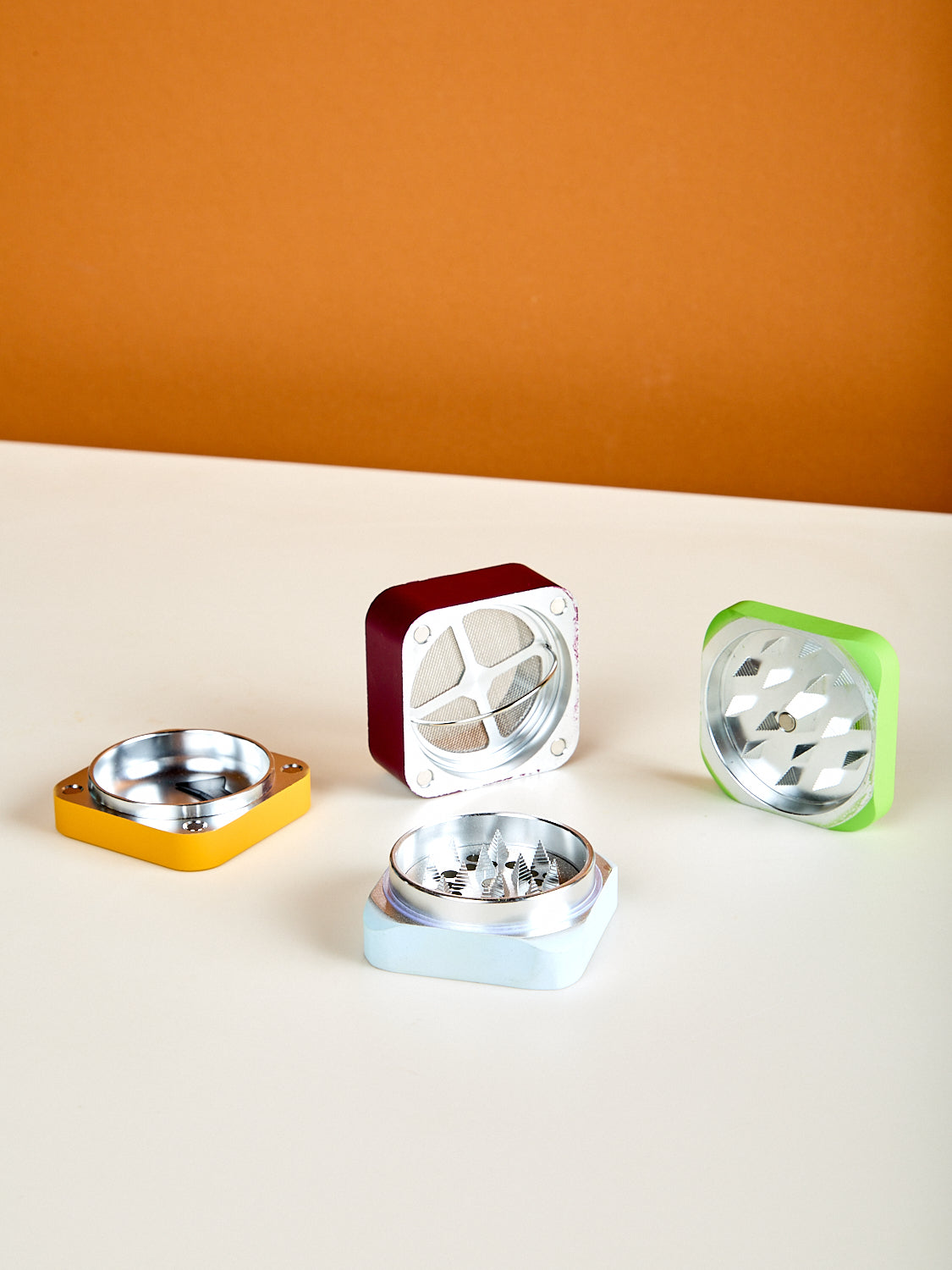 Four metallic square grinder parts with circular centers on a white surface against an orange background