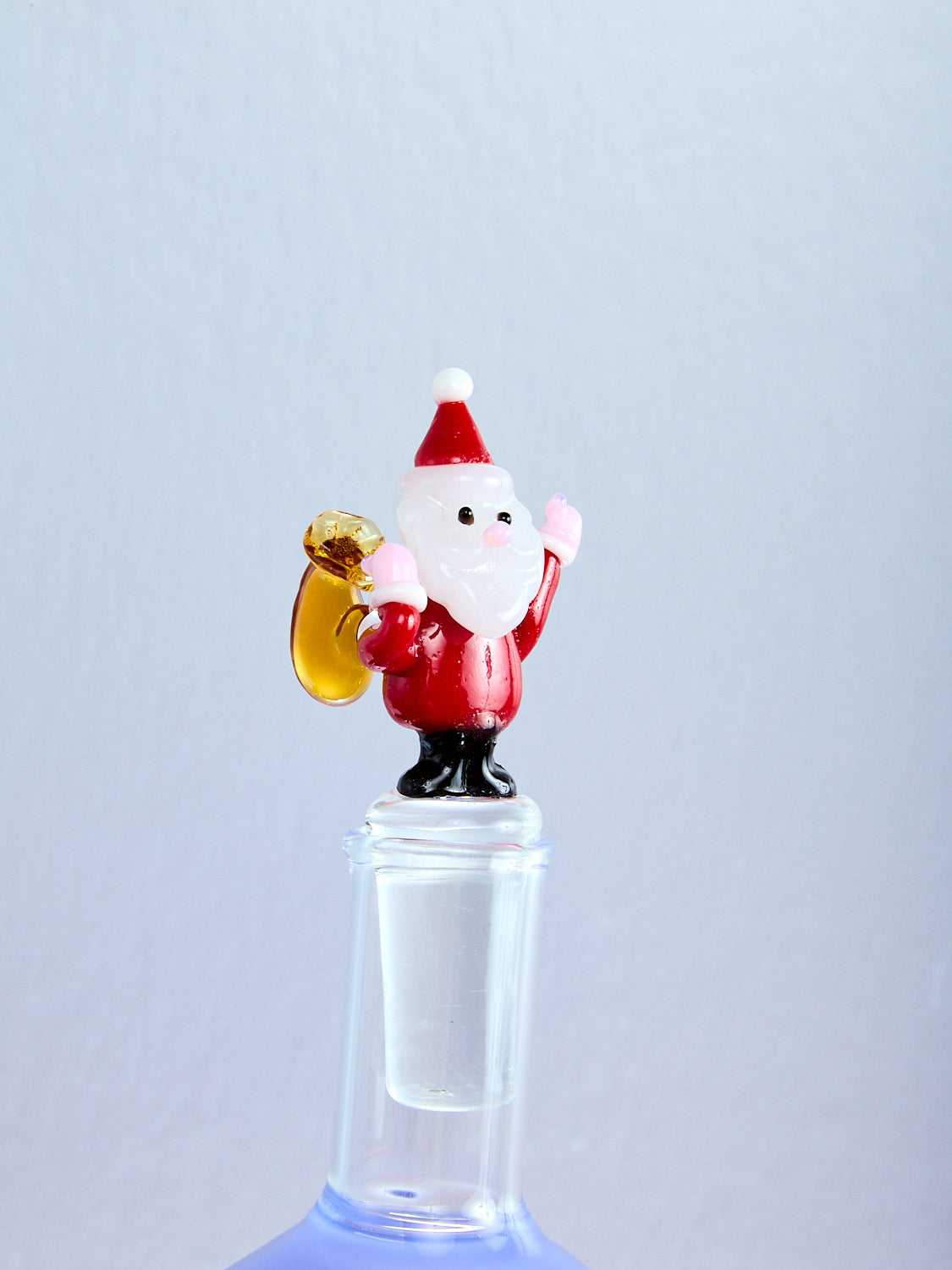 Glass figure of a Santa Claus holding a brown sack on a clear bottle against a light blue background