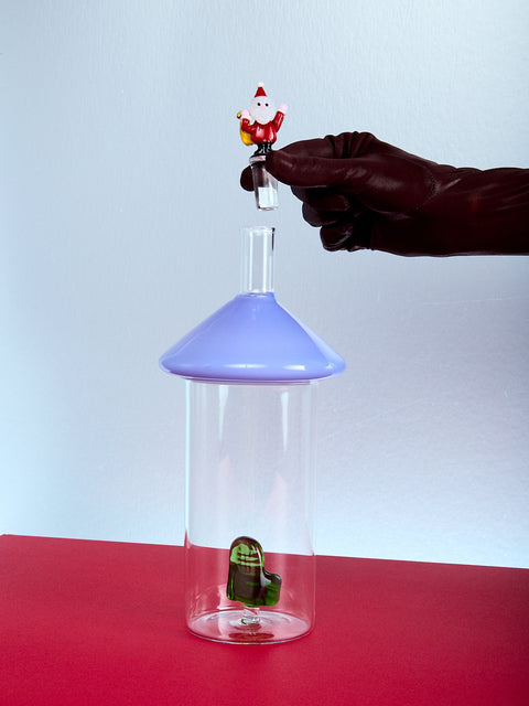 Glass container with a bluish purple top and small santa figurine, held by a gloved hand on a red surface.