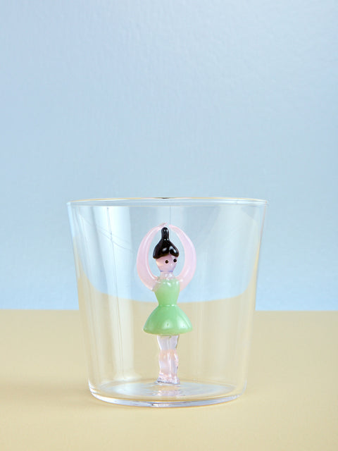 Clear glass with a small figurine of a ballerina in a green dress on a light blue and beige background